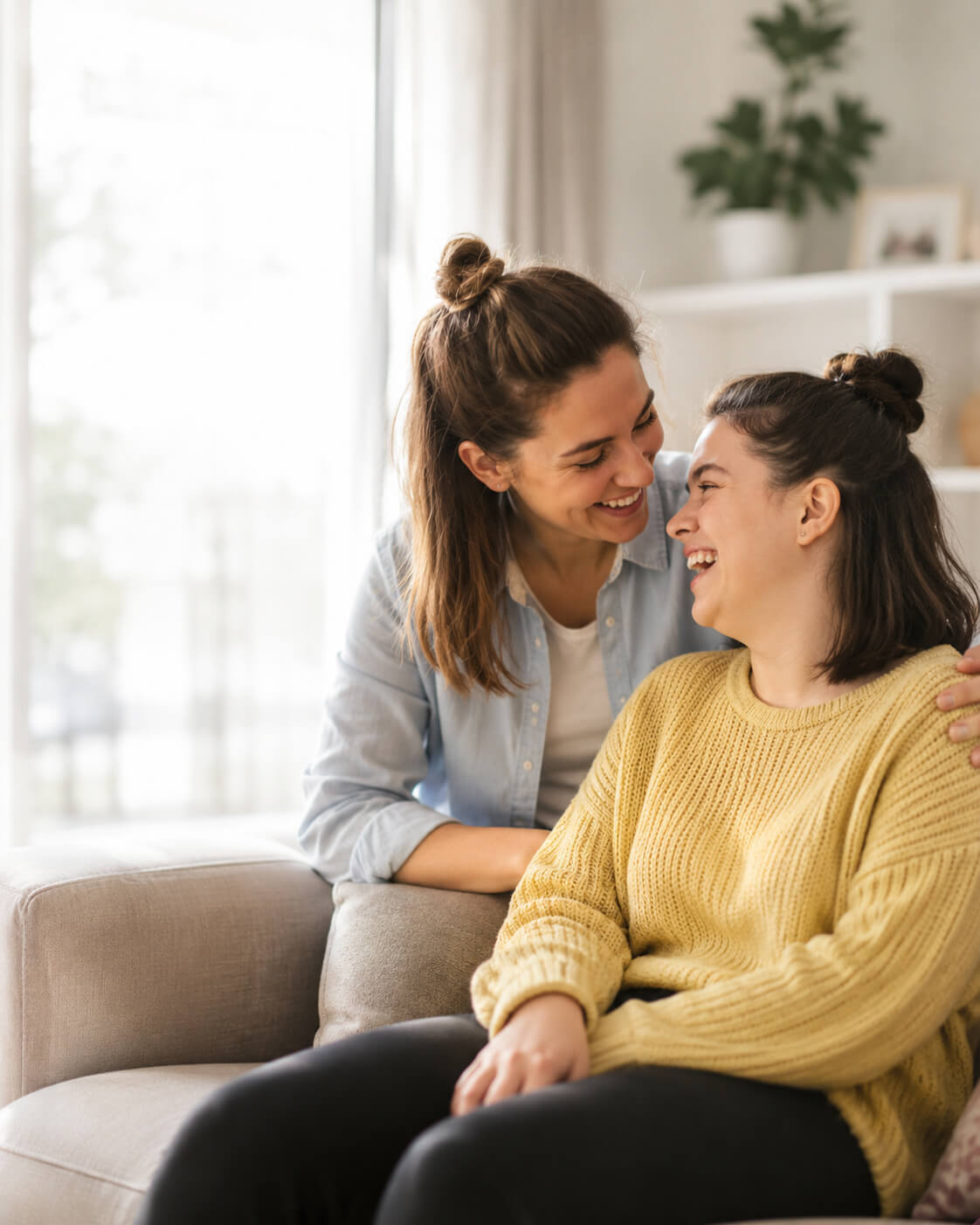 A support worker leaning over a sofa, chatting warmly with a young woman at home, both laughing.
