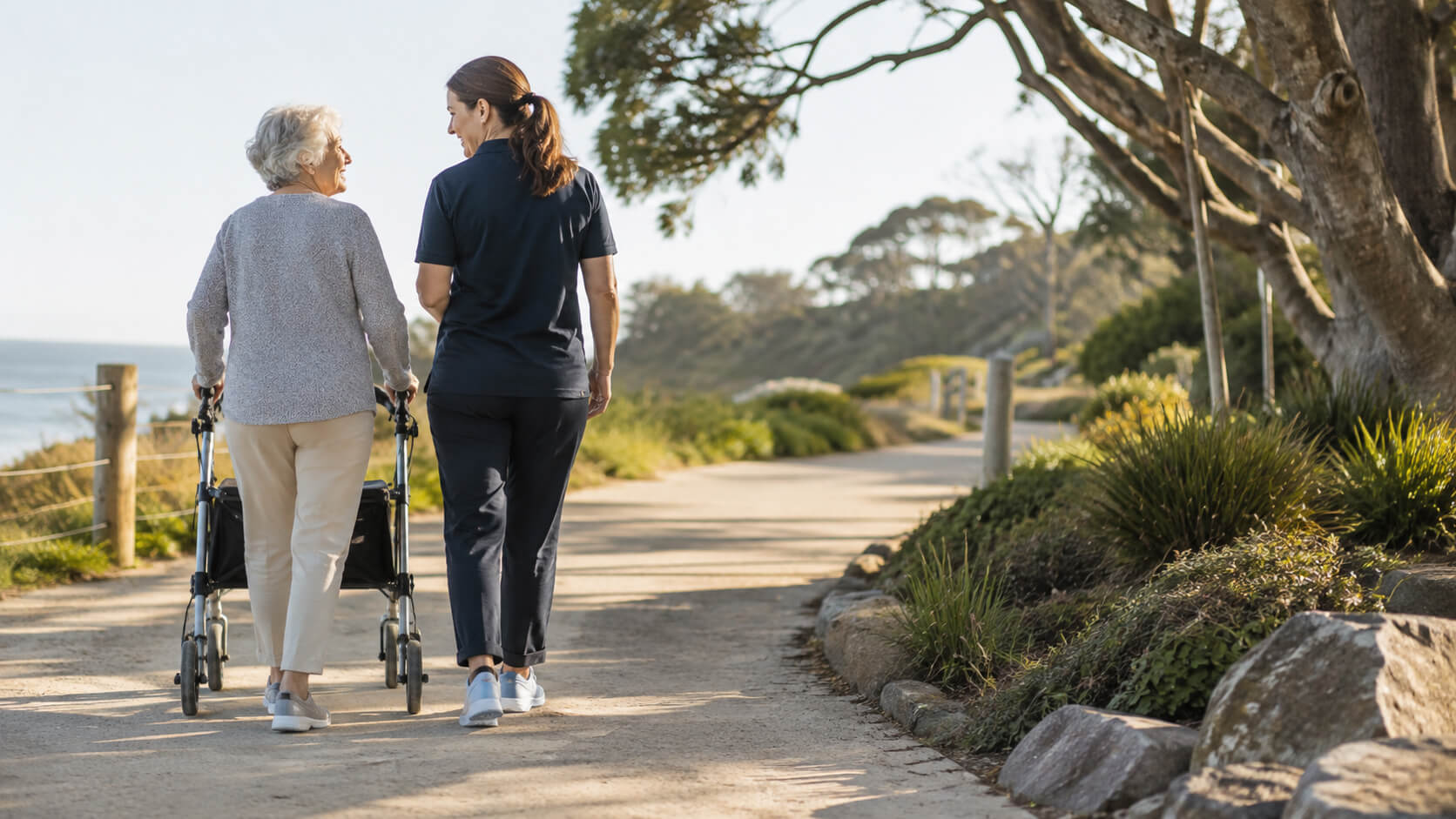 A support worker walking beside an older woman who is using a walking frame on a coastal path.