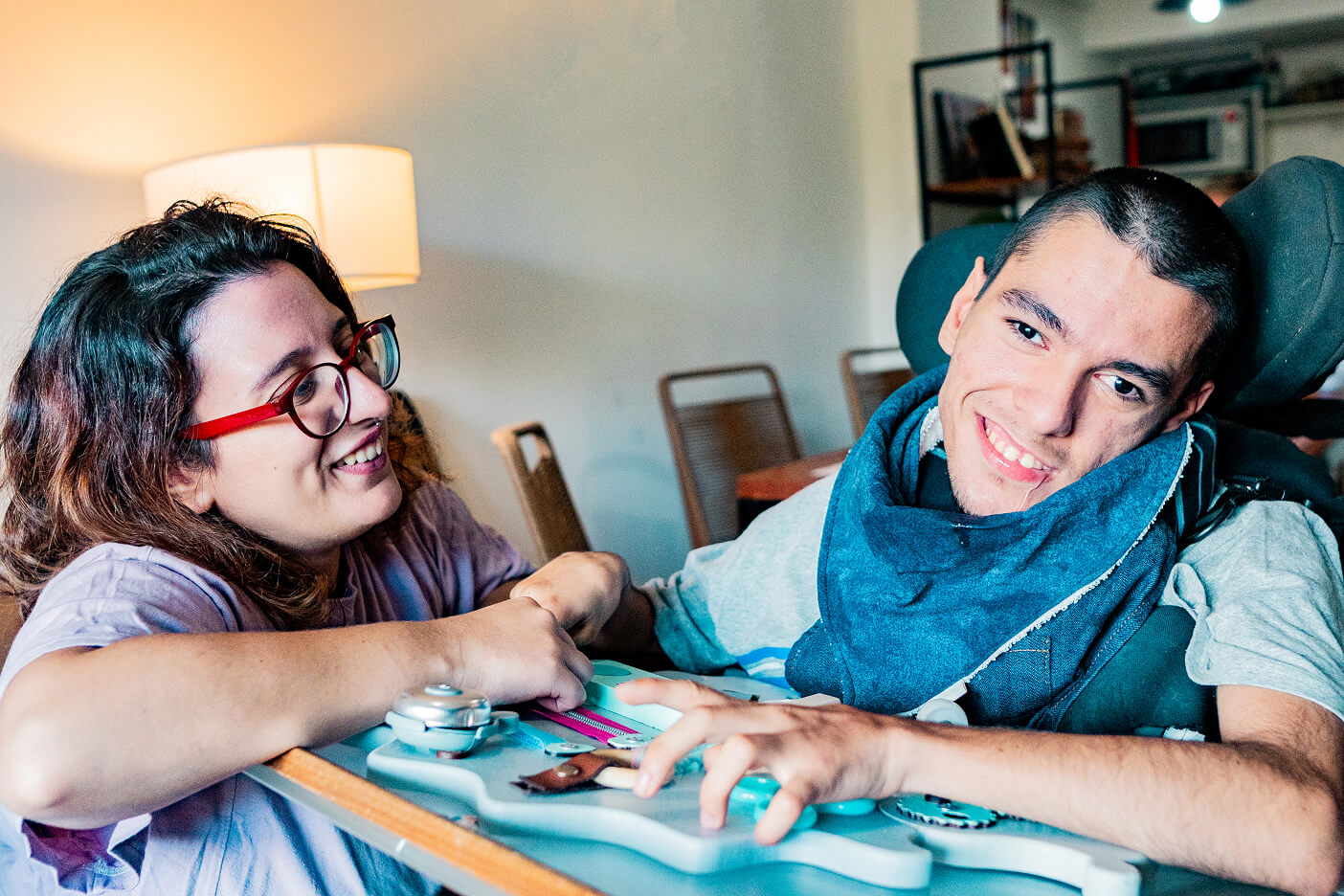 A support worker helping a young man with complex needs use a hands-on activity board at home.