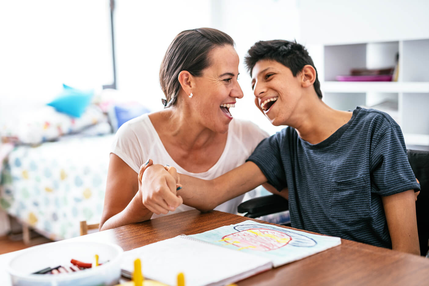 A support worker and a teenage boy laughing over artwork at a kitchen table.