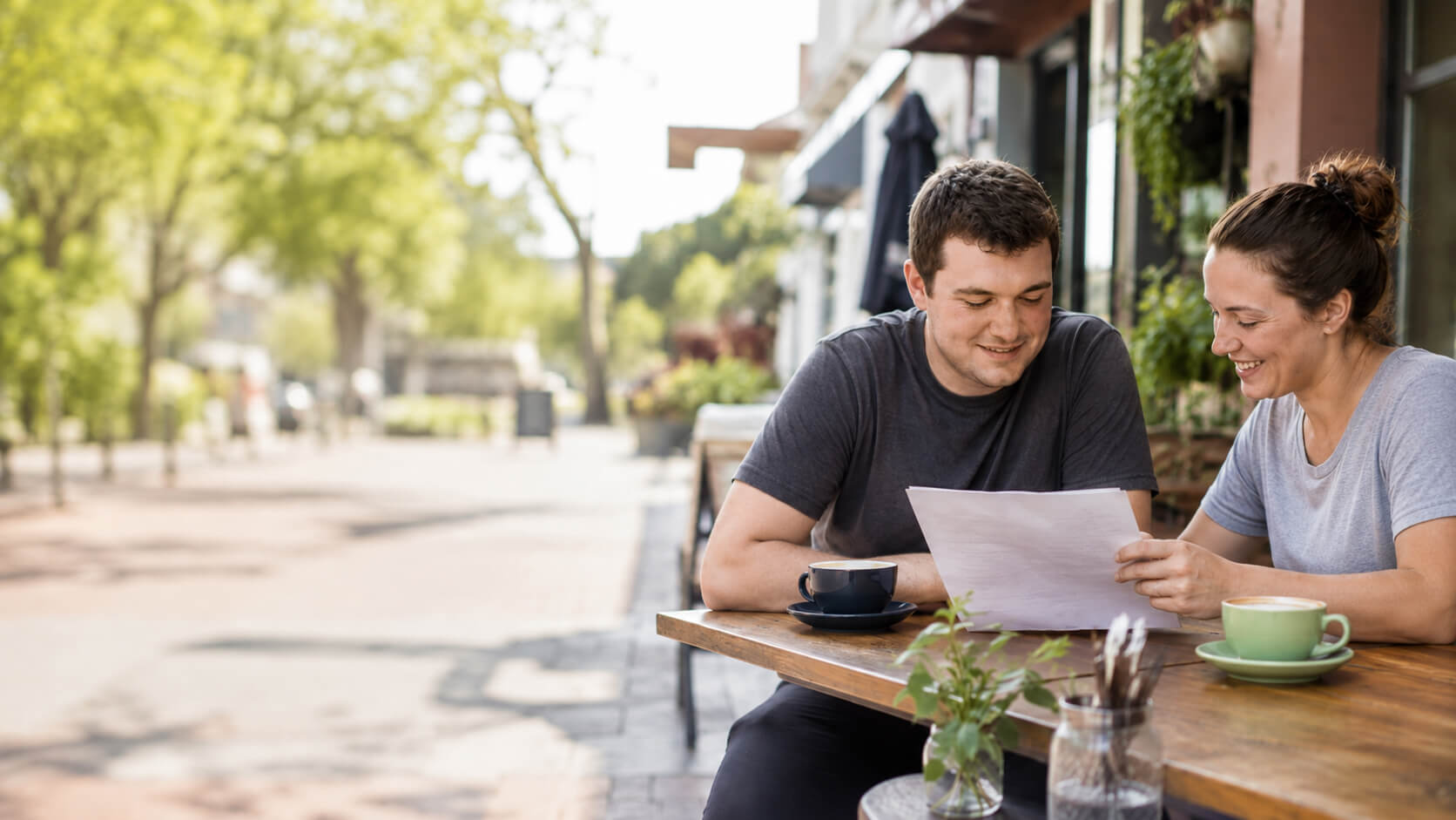 Two people at an outdoor café table, deep in warm conversation over coffee on a sunny street.