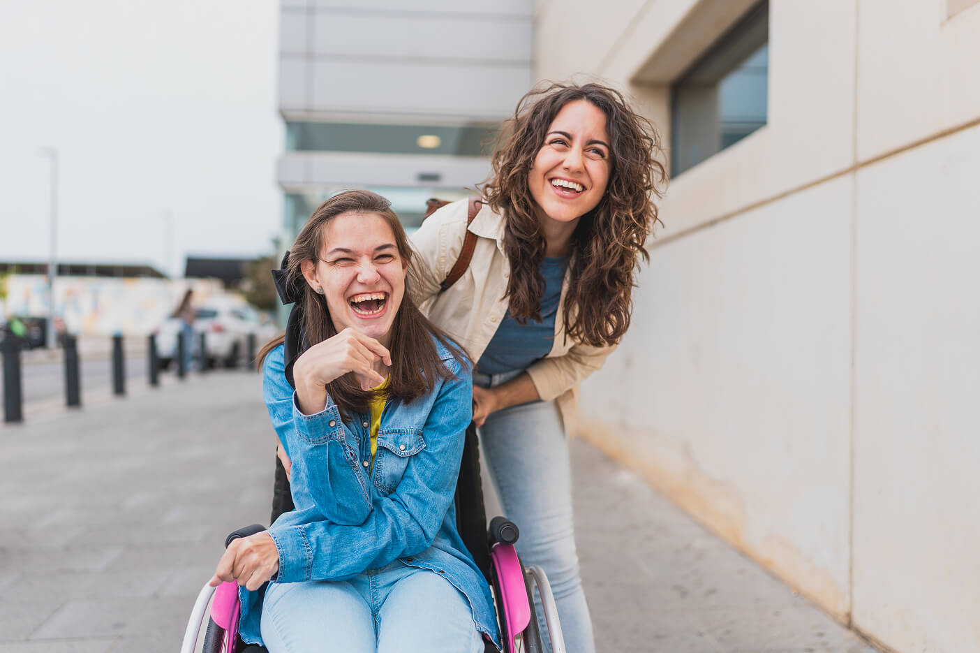 A support worker and a young woman in a wheelchair laughing together on a city street.