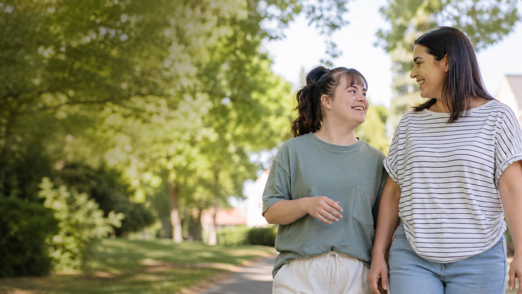 A young woman with Down syndrome and a support worker walking arm in arm through a leafy park.