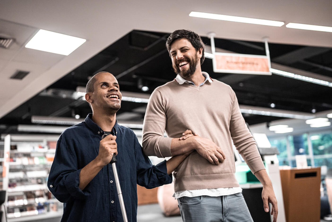 A man using a white cane walking arm in arm with a support worker through a store, both laughing.