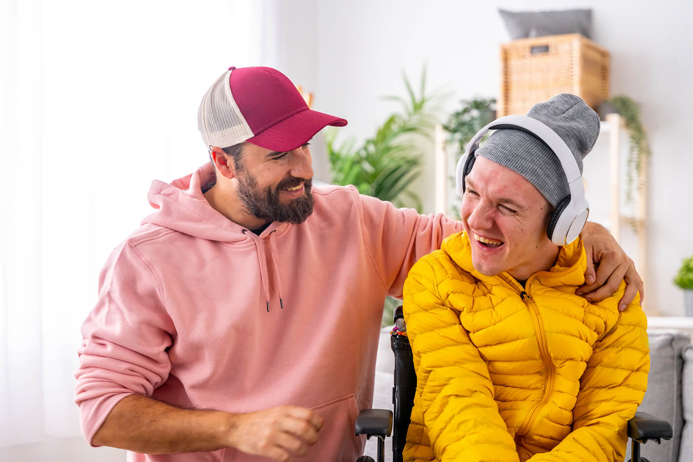 A support worker with his arm around a young man in a wheelchair at home, both laughing.